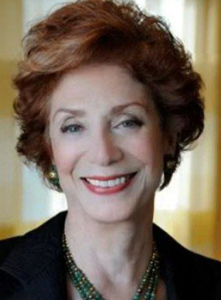A smiling woman with short, curly red hair wearing a black top and a beaded necklace, indoors with a yellow curtain in the background.