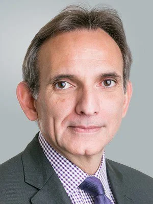 A professional headshot of a middle-aged man with short brown hair, wearing a dark suit, checkered shirt, and tie, against a gray background.