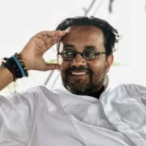 A smiling man with glasses and a beard saluting with his right hand, wearing a white shirt and several bracelets on his arm.