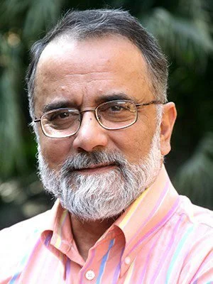 A middle-aged man with glasses, a gray beard, and short hair, wearing a pink and white striped shirt, standing outdoors in front of green foliage.