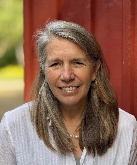 A smiling middle-aged woman with long, gray hair, wearing a white shirt, standing outdoors next to a red wooden structure.