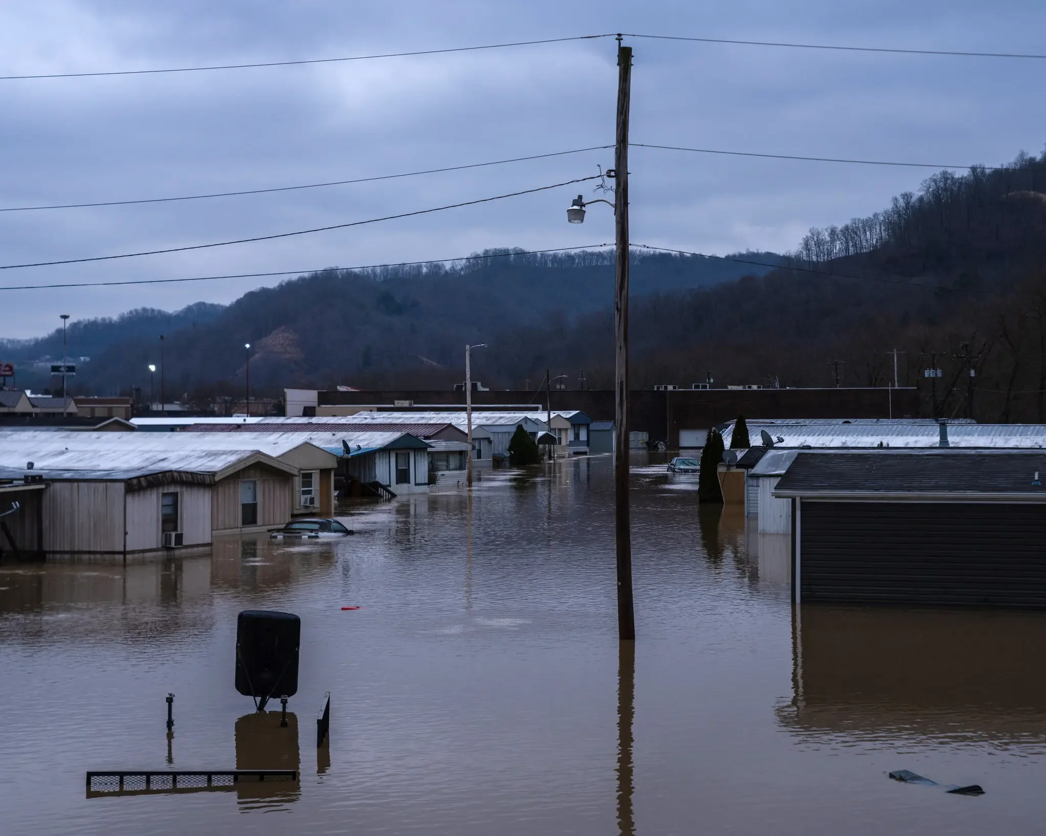 A flooded residential area with water submerging rooftops, utility poles, and trees under a cloudy sky.