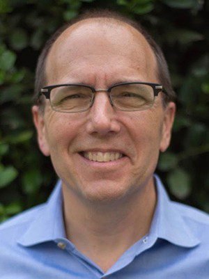 A man with glasses and a blue shirt smiling in front of greenery.