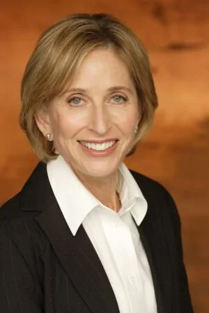 A professional woman with short blonde hair and blue eyes, wearing a black blazer and white shirt, smiling against a wooden background.