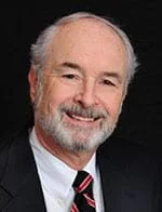 A professional portrait of an older man with a beard, wearing a dark suit, white shirt, and striped tie, smiling at the camera.