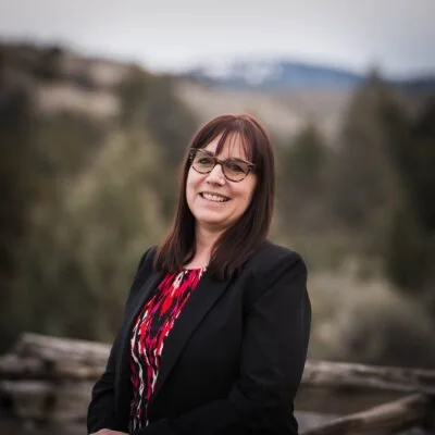 A woman with glasses and straight brown hair smiling outdoors in a scenic natural setting with trees and mountains in the background.