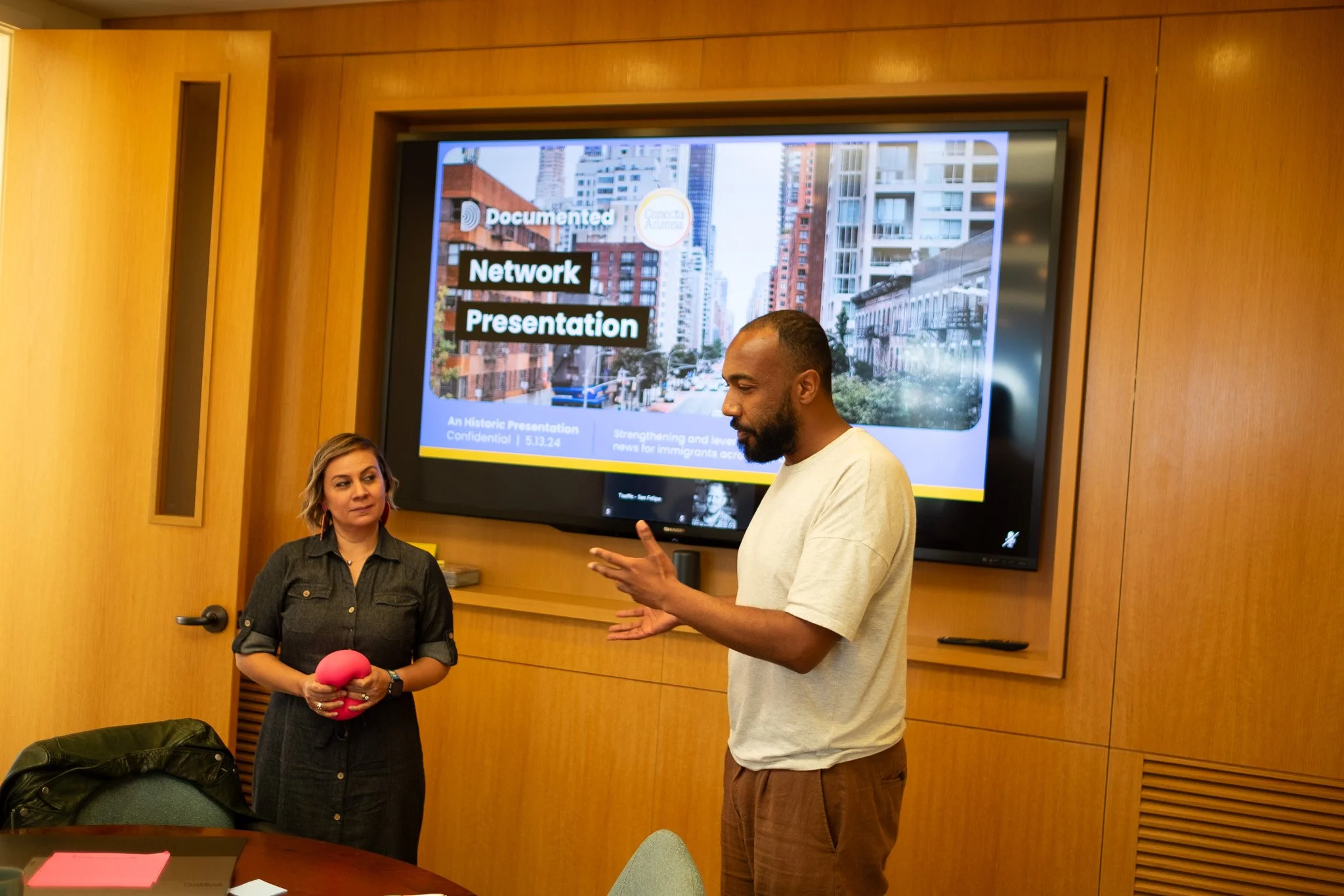 Two people are standing in a room with wooden walls. Behind them is a large screen displaying a cityscape with tall buildings and text that reads 'Documented Network Presentation.' The woman on the left is holding a pink plush object, and the man on the right is speaking and gesturing with his hand.