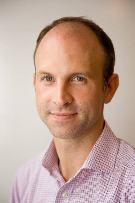 A man with light skin, short light brown hair, and blue eyes looking at the camera, wearing a pink checkered shirt against a plain neutral background.