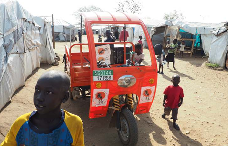 A red three-wheeled vehicle with cartoon character decals on the front, surrounded by children and tents in a makeshift outdoor setting.