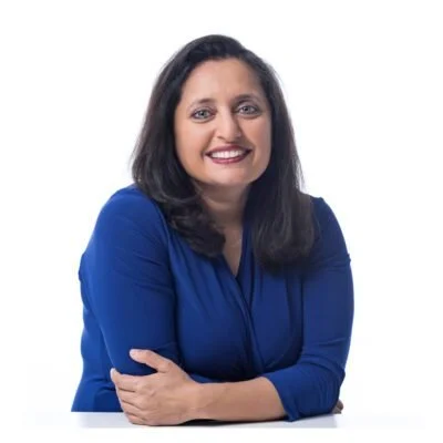 A woman with dark hair and a bright smile wearing a blue blazer, sitting with her arms crossed against a white background.
