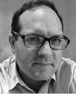 A black-and-white photo of a man wearing glasses and a collared shirt, looking at the camera.