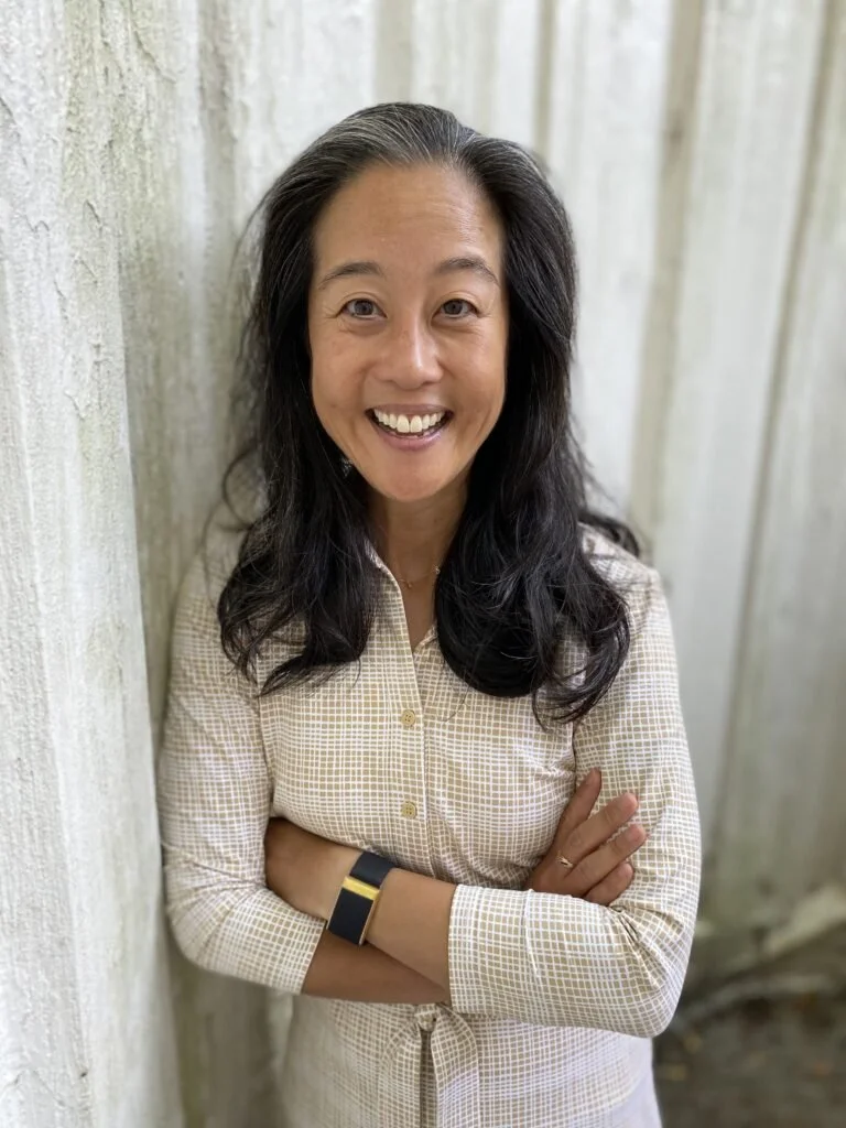 A woman with long dark hair smiling and crossing her arms, standing against a wooden fence.