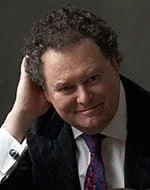 Portrait of a man with curly dark hair, wearing a dark blazer and a patterned tie, smiling.