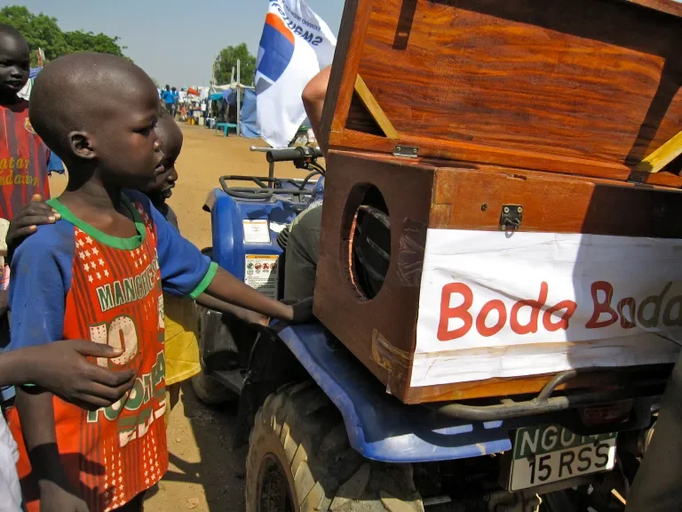 Young boy in a red and orange sports jersey standing next to a wooden box with a speaker inside, placed on a blue vehicle at an outdoor event, with people and tents in the background.