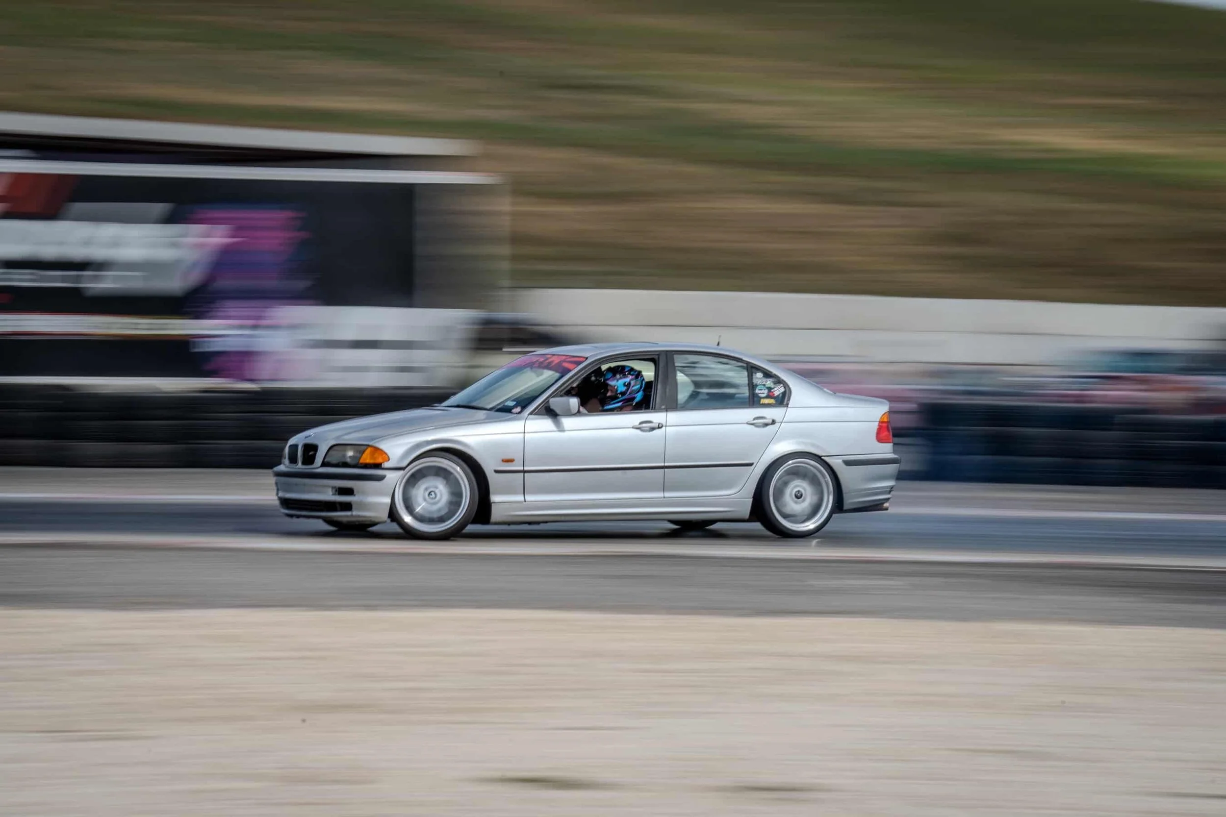 Silver sports car with a driver in racing gear speeding on a racetrack with a blurred background.