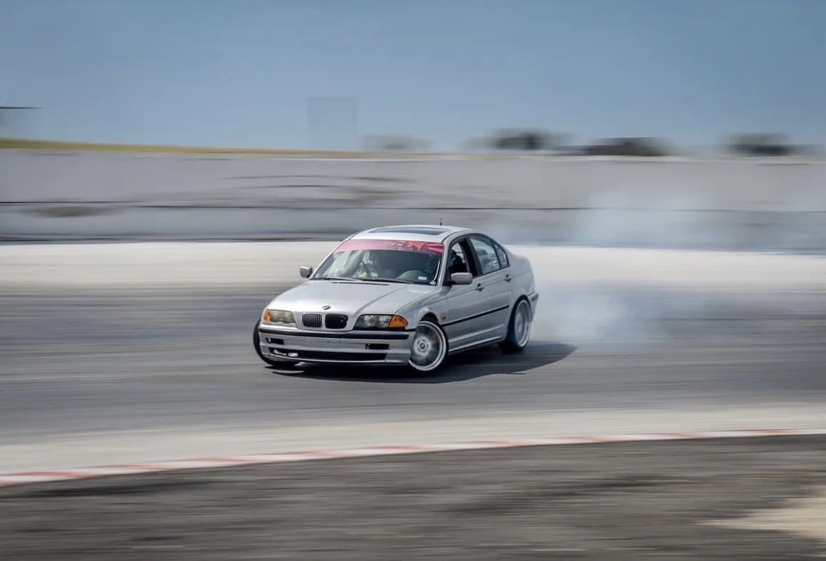 A silver BMW drifting on a race track, kicking up smoke from the tires.