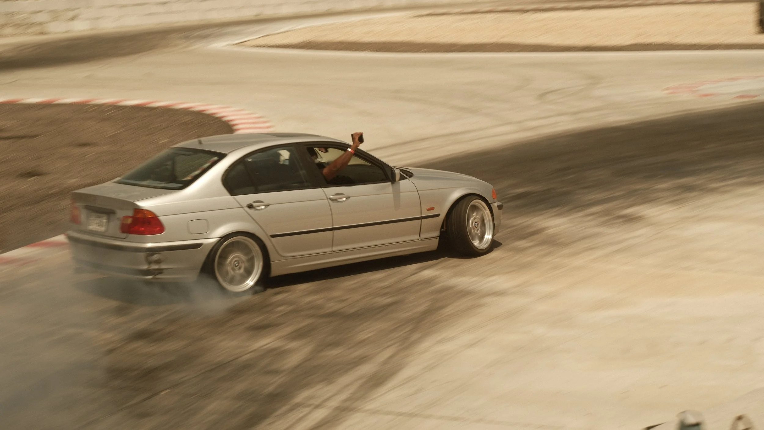 A silver car on a race track with a person inside raising a fist through the window during a drift action