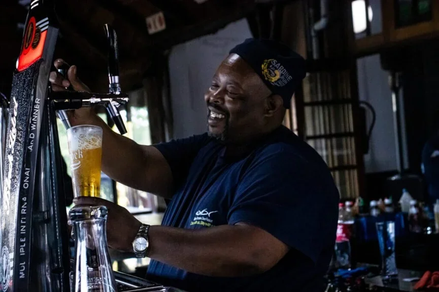 A man smiling while pouring a beer from a tap at a bar or pub.