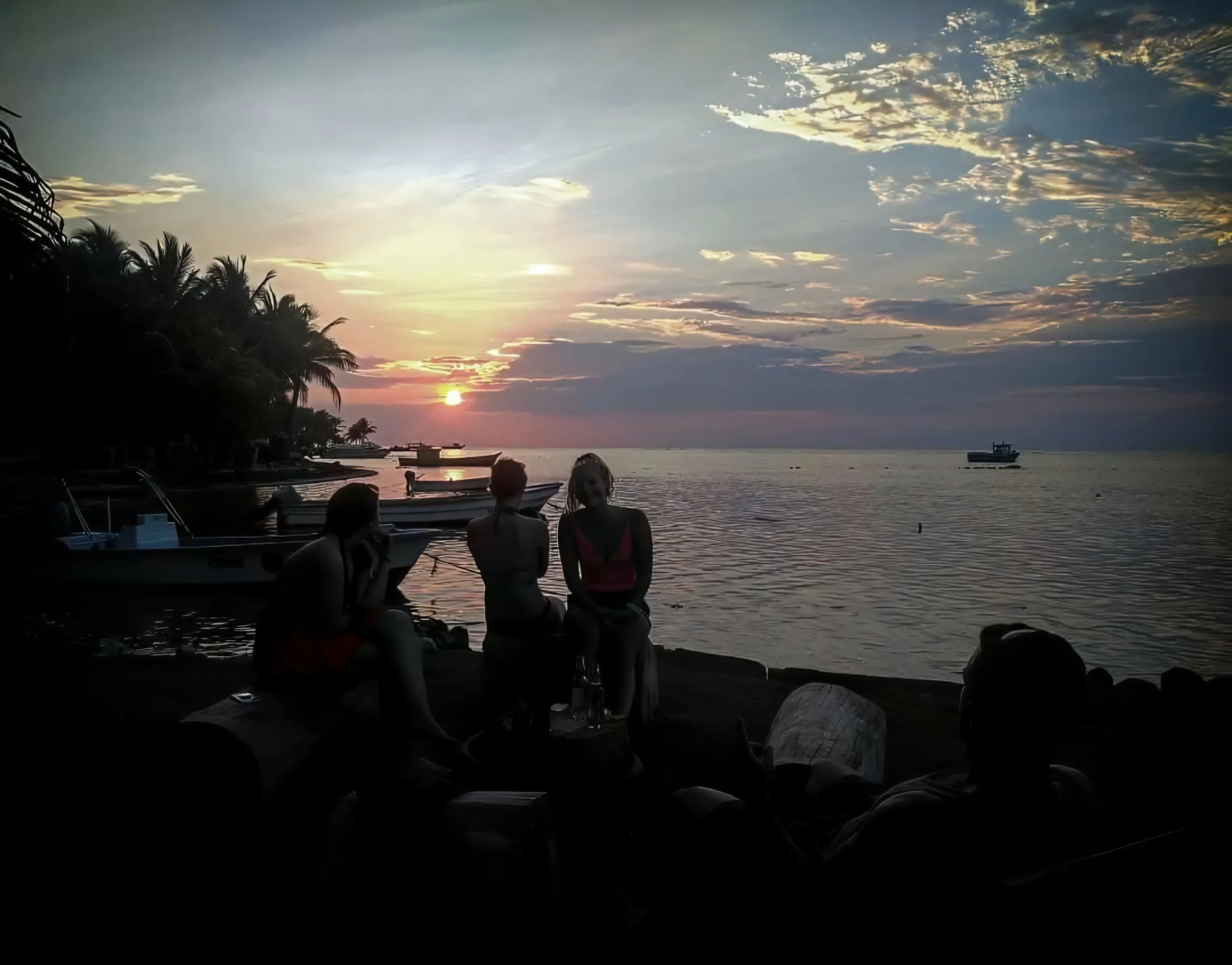 People sitting on the beach during sunset, with boats on the water and palm trees along the shoreline, some clouds in the sky.