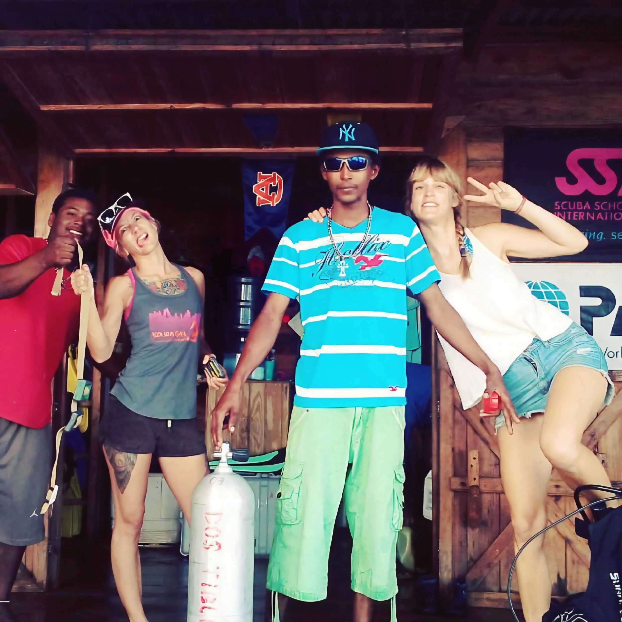 Four young people posing in front of wooden background with diving-related banners. One person is wearing sunglasses, a black cap with the New York Yankees logo, and a light blue and white striped shirt. The others are smiling and making peace signs.