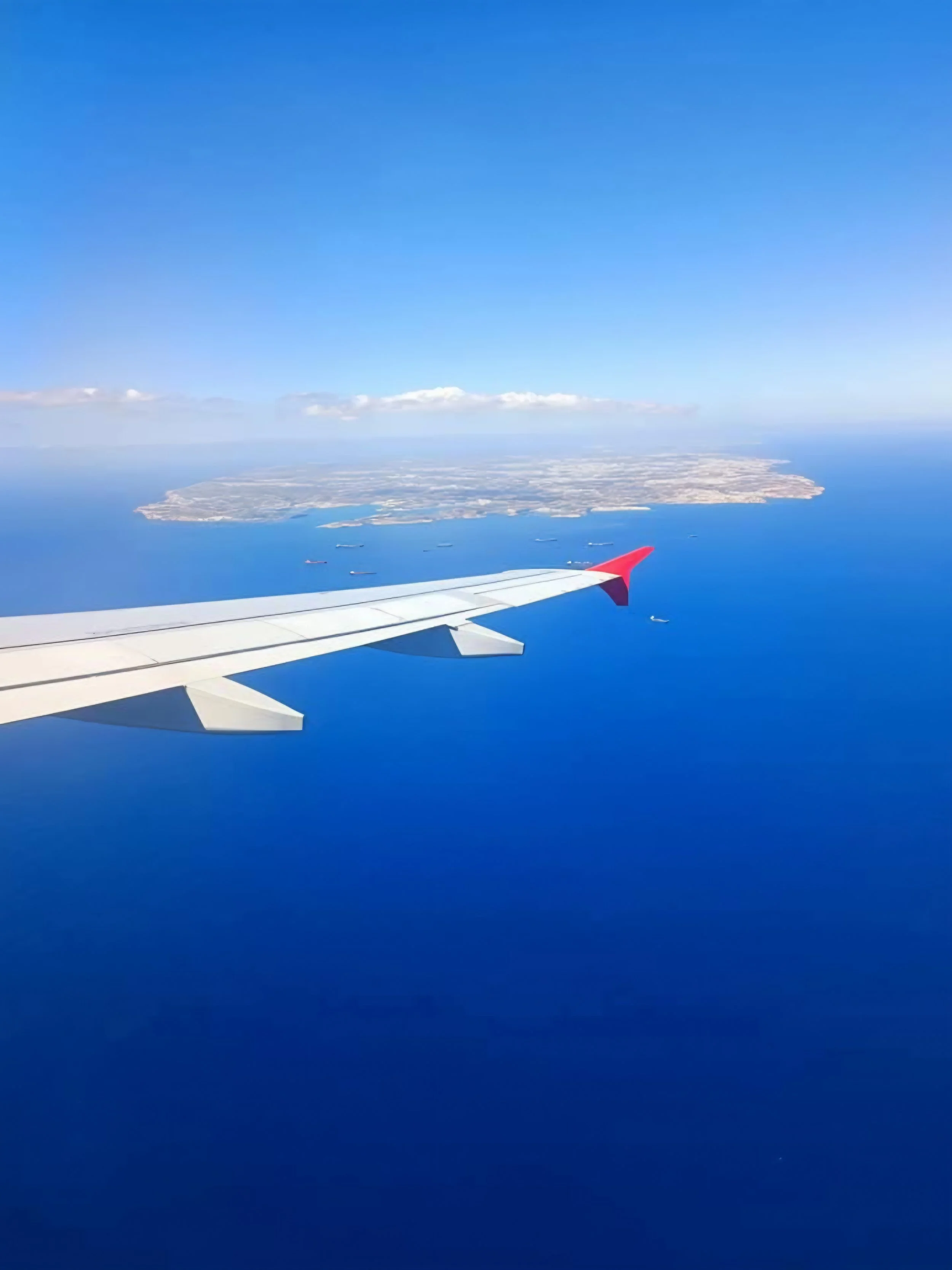 A view from an airplane window showing a clear blue sky, a distant coastline, and ships in the ocean below.