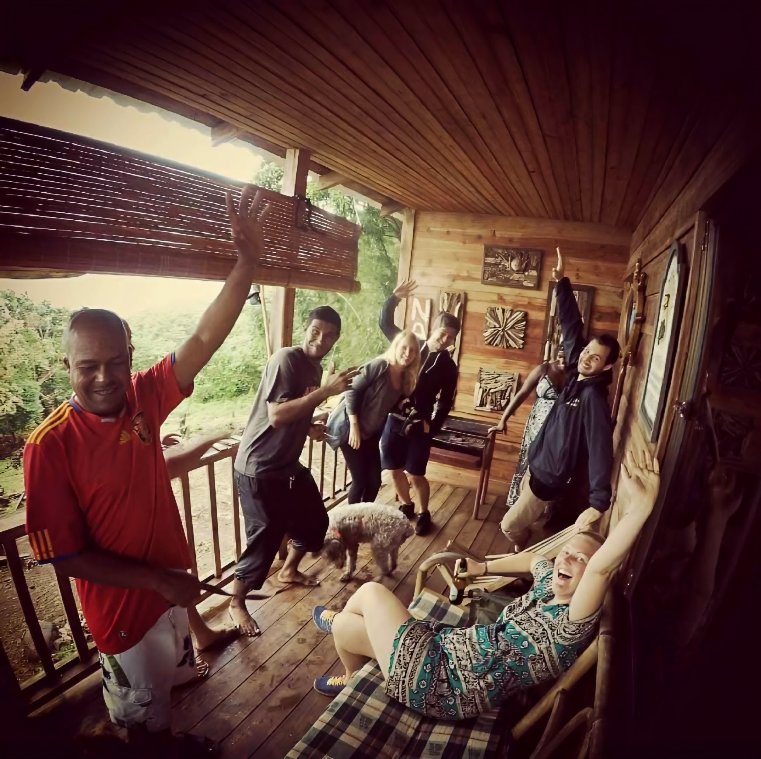 Group of people inside a wooden cabin, some sitting and some standing, with a person raising their hand, and a person in a red shirt in the foreground, surrounded by greenery outside.