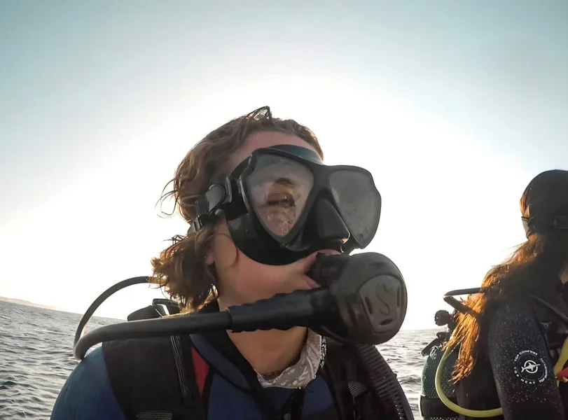 A woman wearing diving gear, including a mask and regulator, on a boat or near water with the ocean in the background.