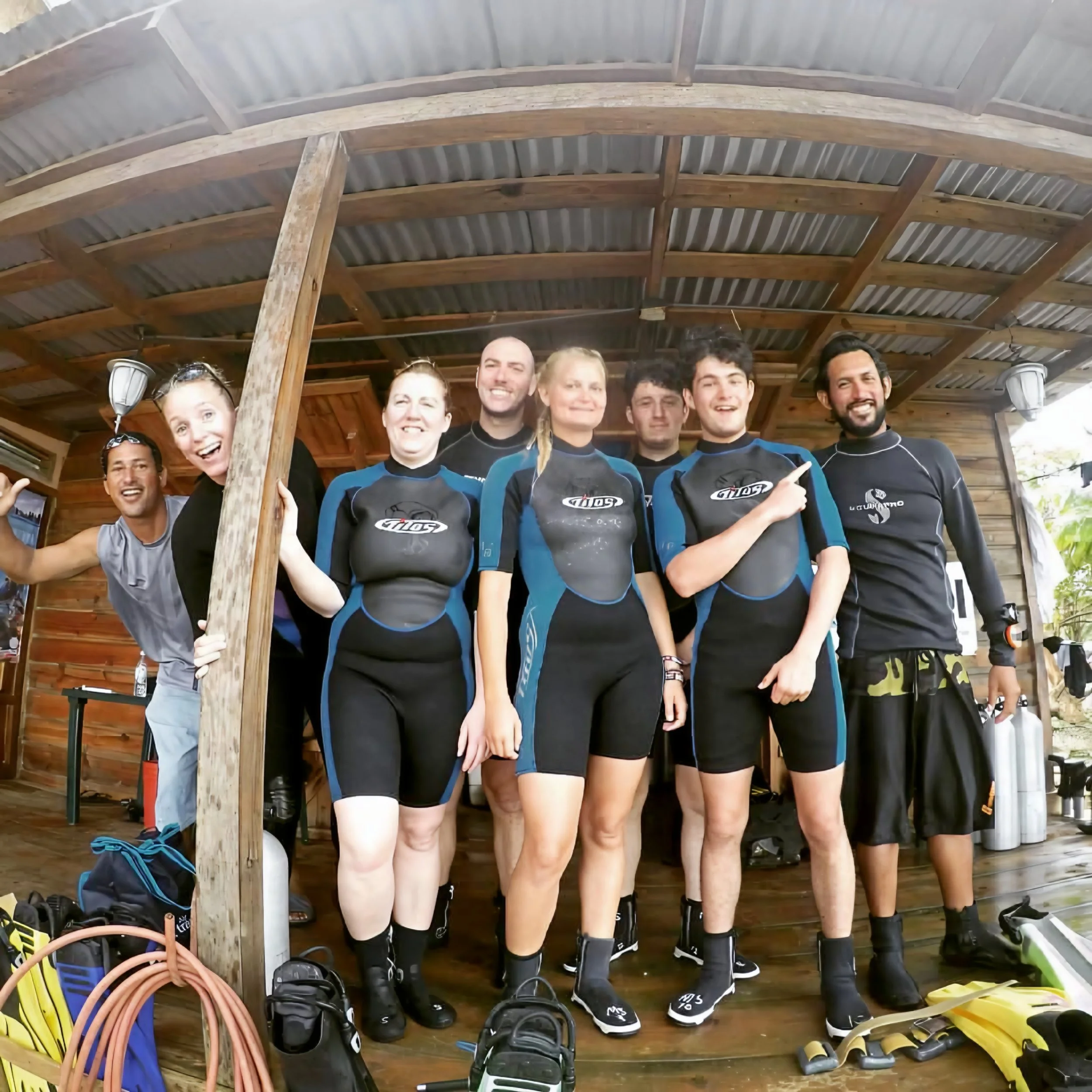 Group of people standing under a wooden roof, smiling and posing for a photo, with outdoor gear such as boots and climbing equipment on the ground in front of them.