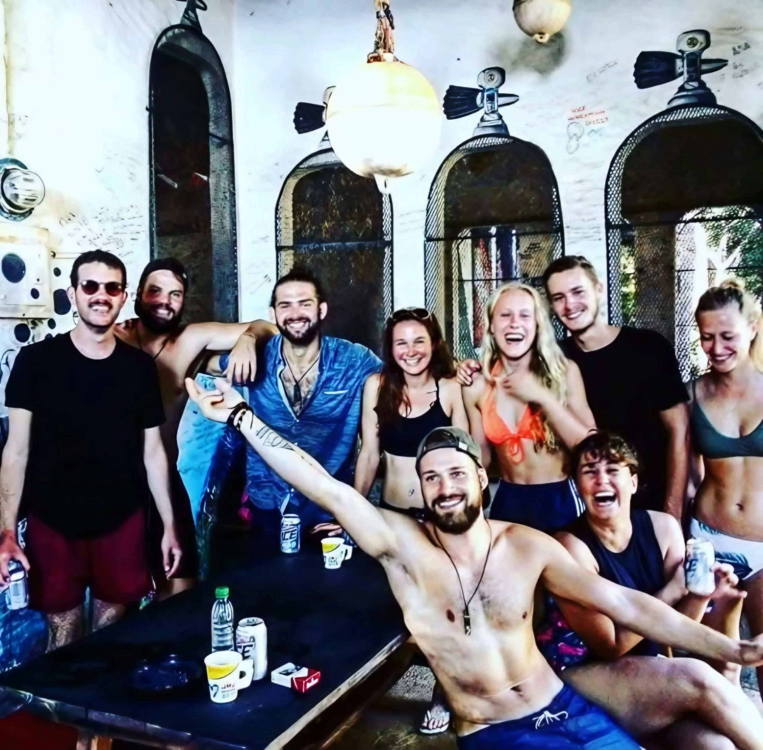 Group of smiling young adults at a party, some shirtless, standing around a table with drinks, under a covered outdoor area.