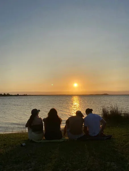 Four people sitting on the grass by the water during sunset.