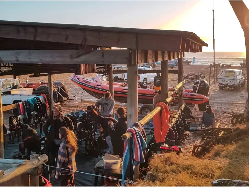 People near a beachside rental hut with boats and watercraft, during sunset.