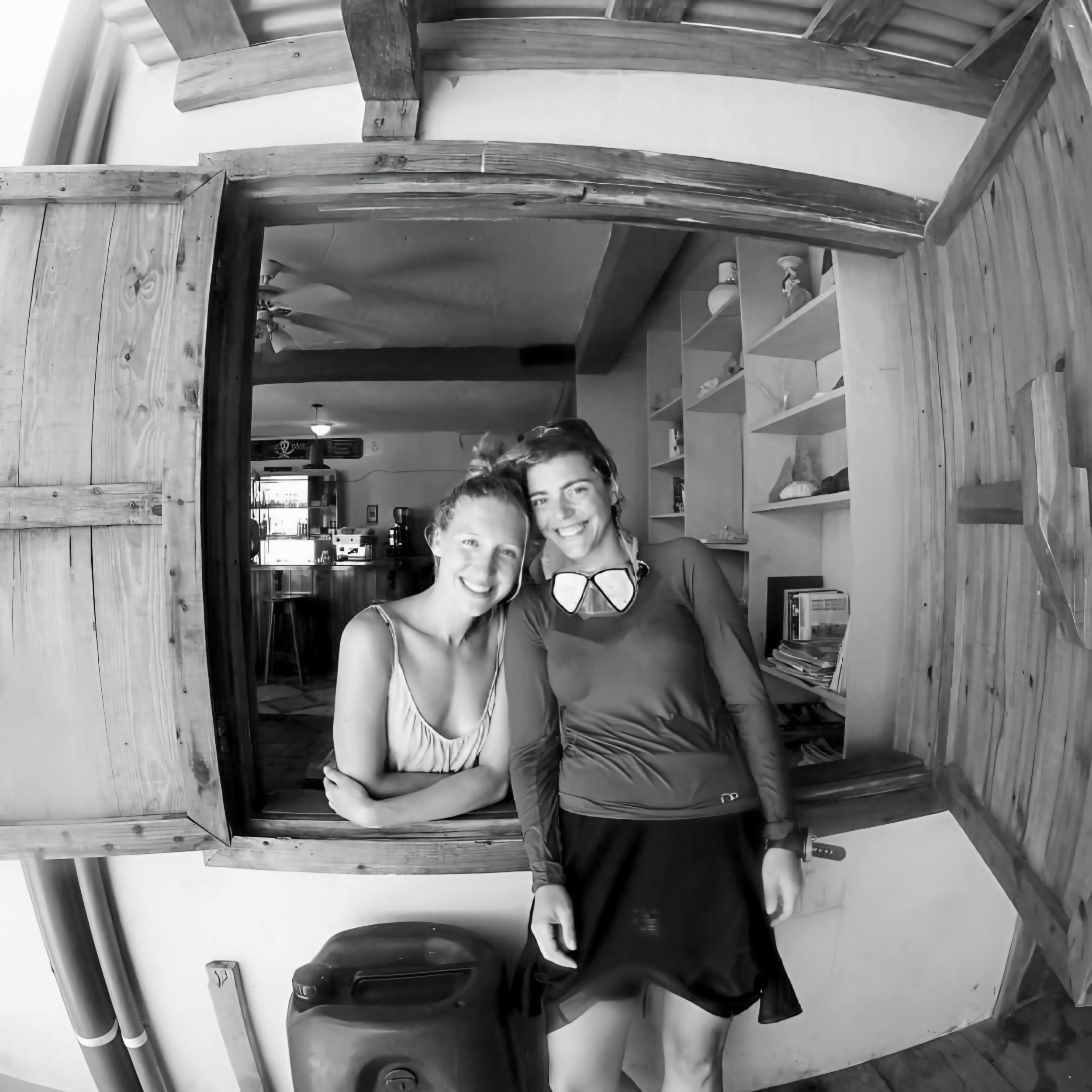 Two people, a woman and a child, smiling in front of a mirror in a cozy room, with wooden beams and furniture, including a cabinet with shelves holding small objects, and a ceiling fan visible in the reflection.