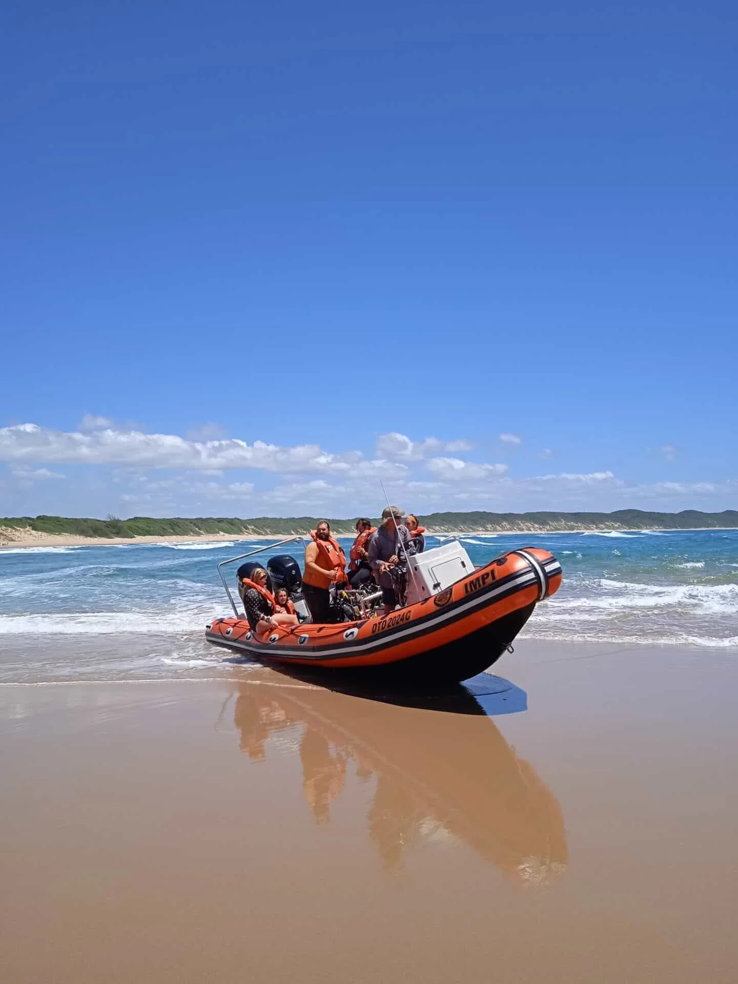 A group of people in life jackets on an inflatable motorboat near the beach shore, with the ocean and cloudy sky in the background.