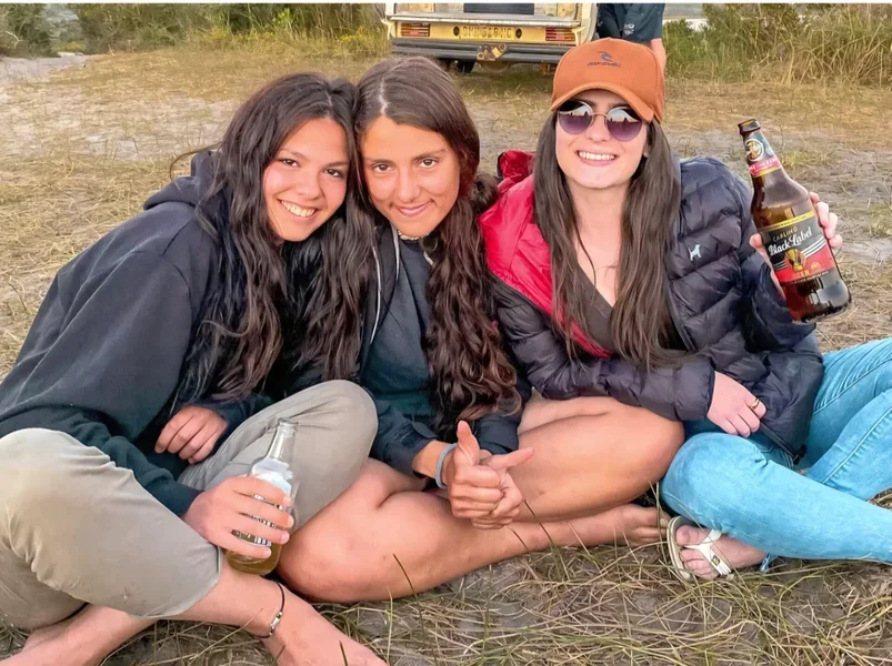 Three young women sitting close together on the ground outdoors, smiling at the camera. Two are holding bottles of beer, and one is giving a thumbs-up. They are dressed casually with jackets and hats, and there is a vehicle in the background.
