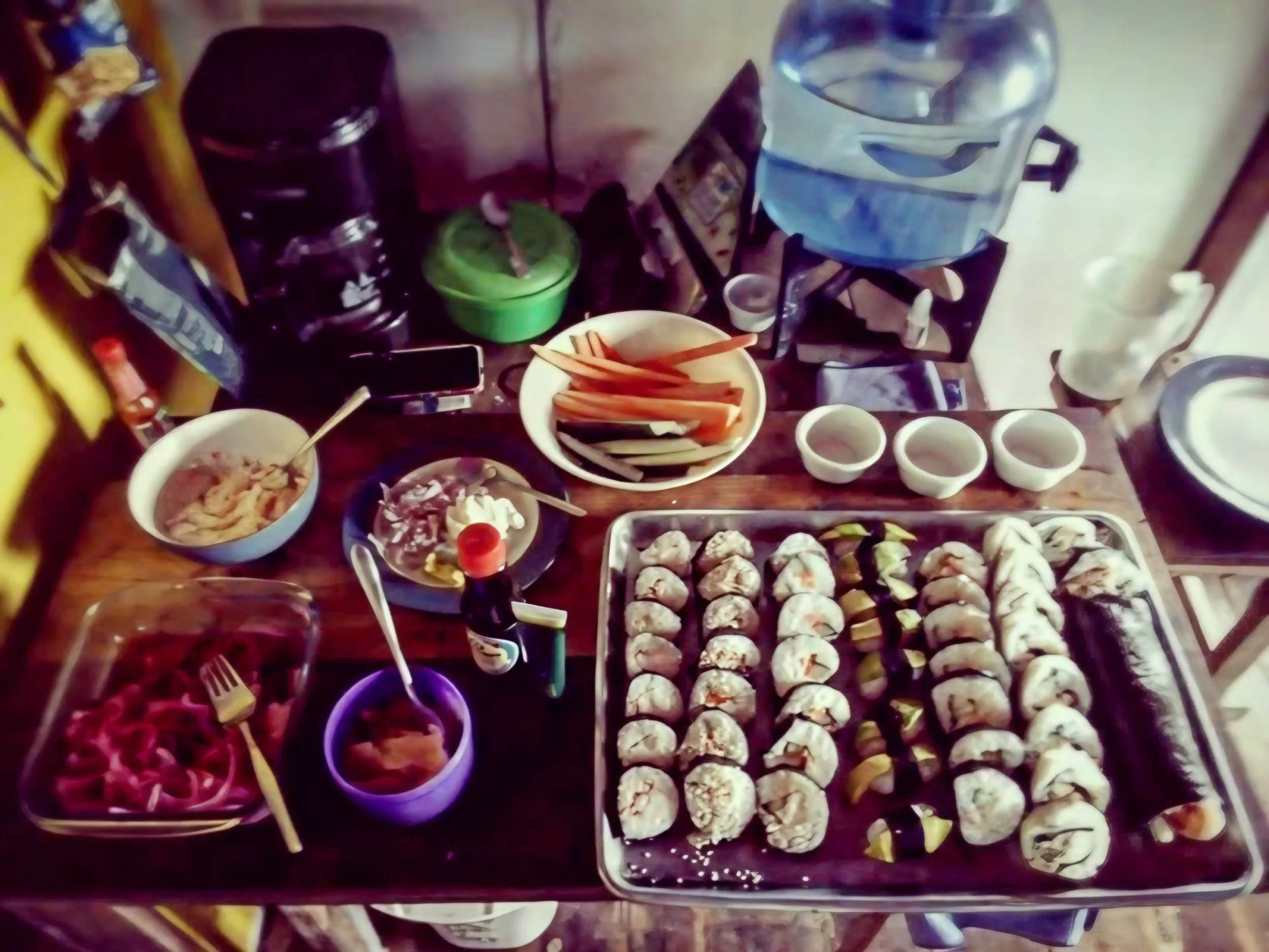 A table filled with various types of sushi, dipping sauces, a large water dispenser, a bowl of fruit with slices of vegetables, and other condiments and food items.