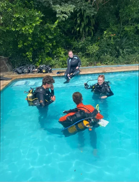 Four people practicing scuba diving in a swimming pool, with lush greenery in the background.