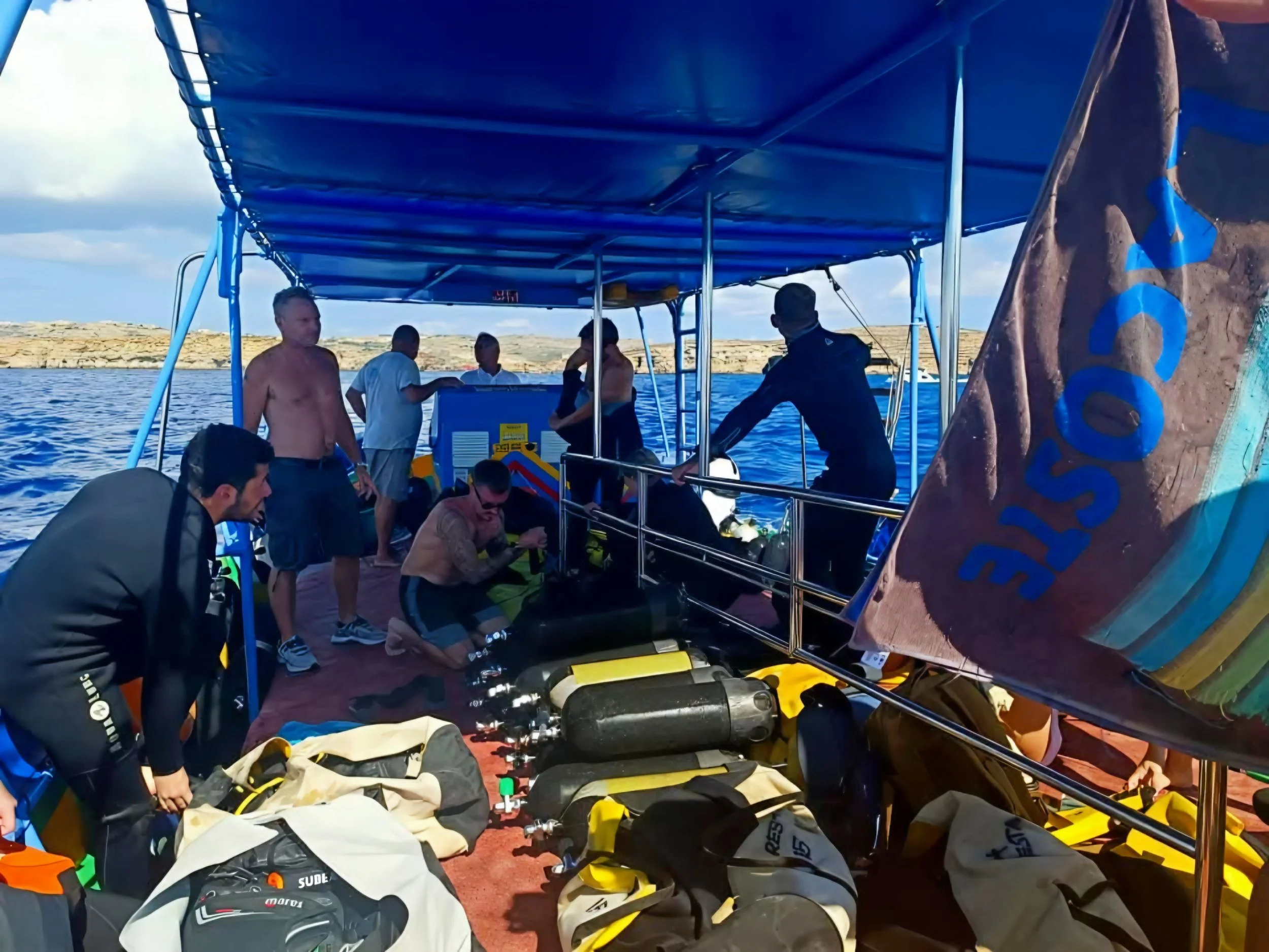 People preparing diving or snorkeling gear under a blue canopy on a boat, with helmets, fins, and other equipment on the ground, and rocky landscape in the background.