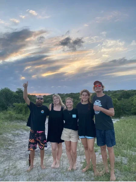 A group of five people standing outdoors on a grassy area with trees and a cloudy sky at sunset in the background.