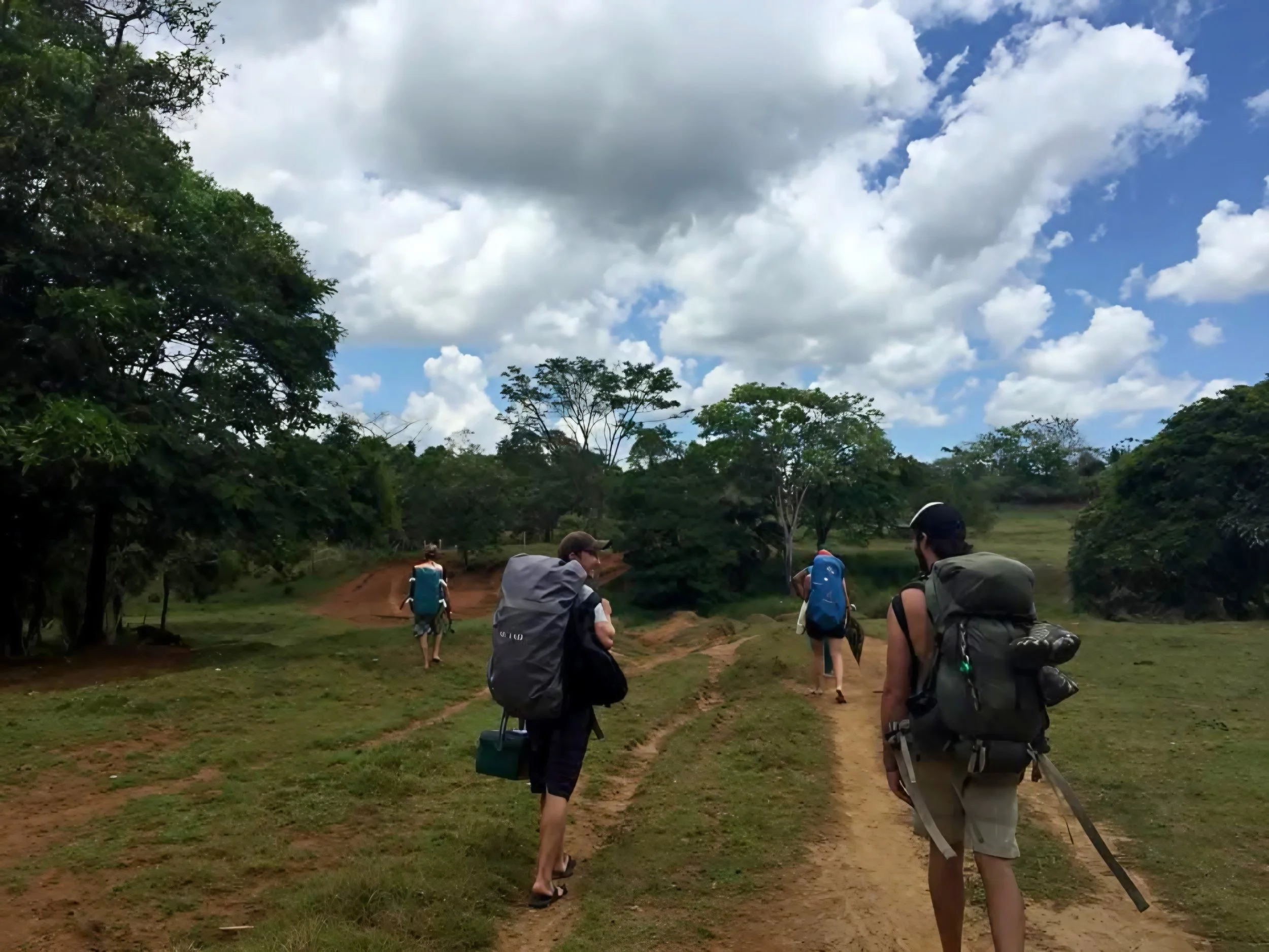 Group of hikers walking along a dirt trail through a grassy area surrounded by trees under a partly cloudy sky.