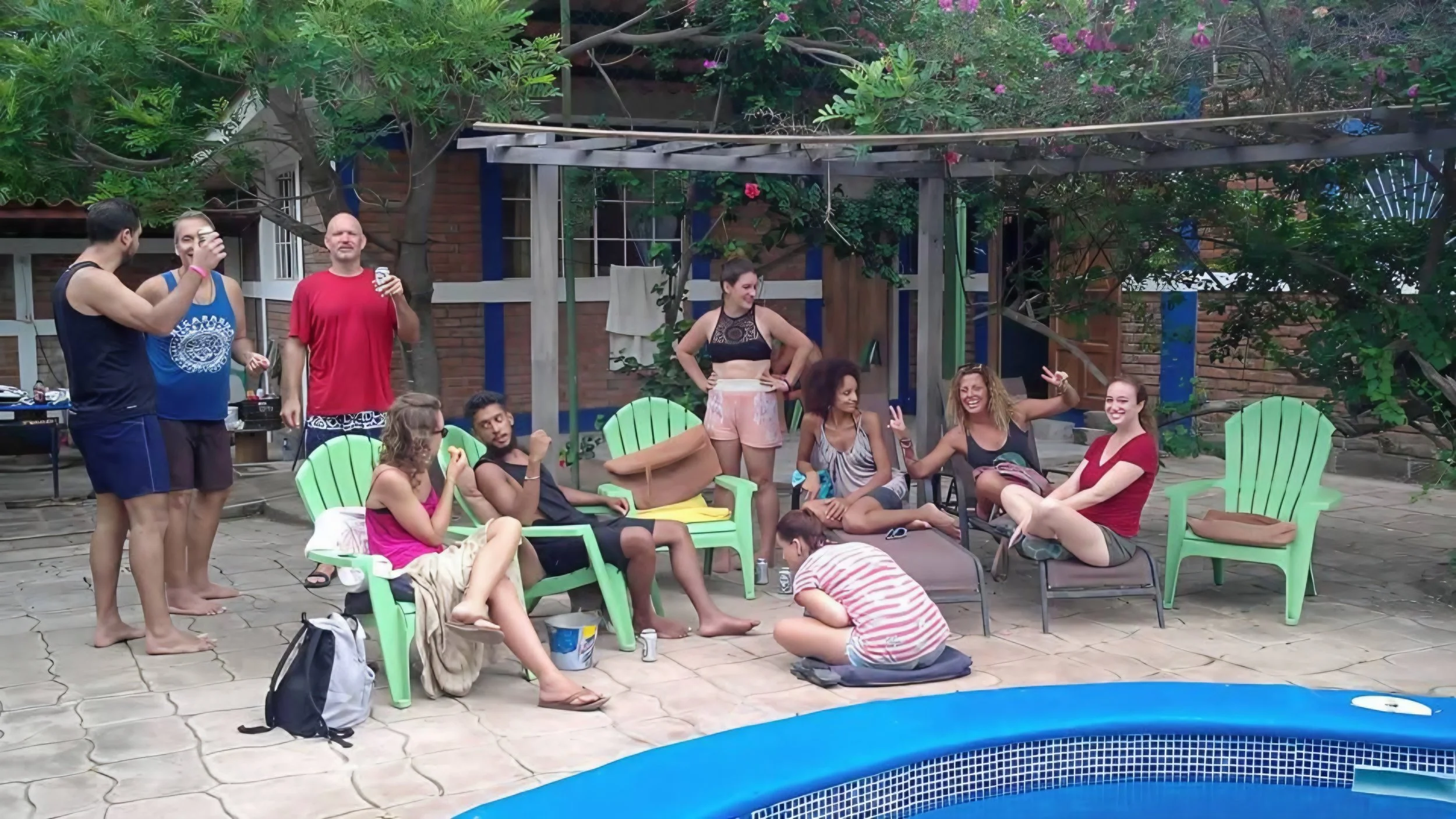 A group of friends gathered outside around a pool, sitting on green Adirondack chairs, engaging in conversation, with some standing and some seated, surrounded by trees and a brick building.
