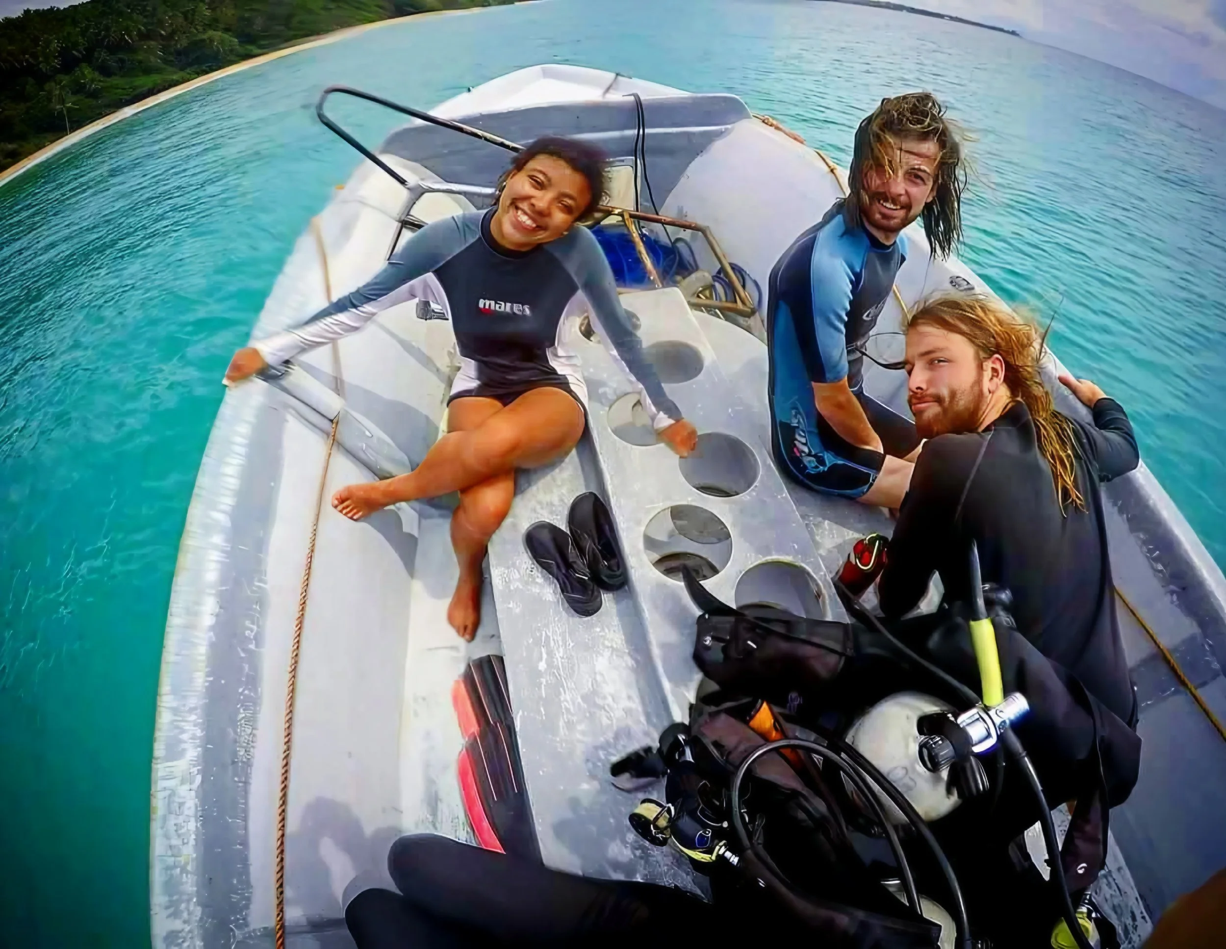 Three people smiling on a small boat in clear blue water, with a lush green coastline in the distance.