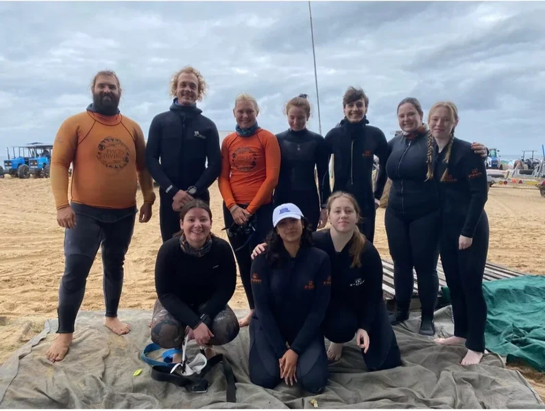 Group of nine people, some in wetsuits, at the beach on a cloudy day, with vehicles and equipment in the background.