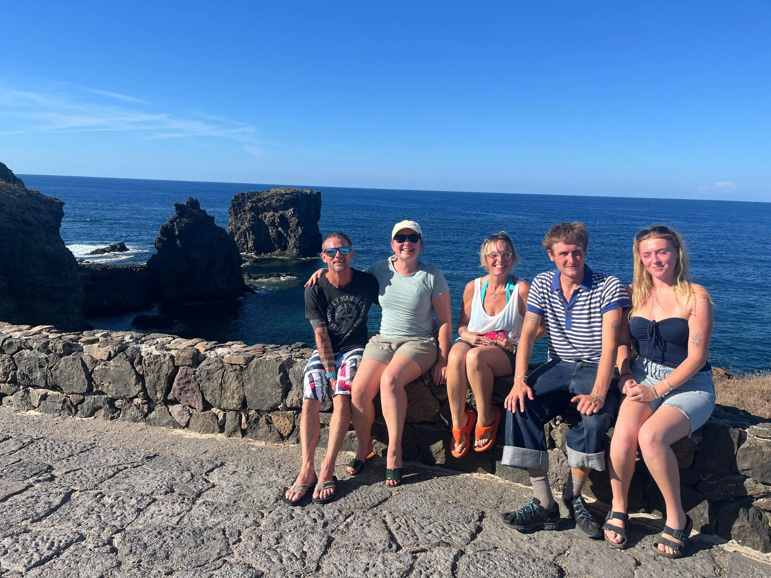 A group of six people sitting on a stone wall at the edge of the ocean on a sunny day, with rocky cliffs and the sea in the background.