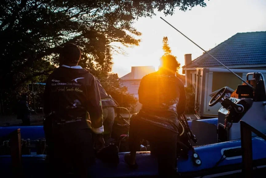 Two people preparing a boat for seafaring at sunset, with trees in the background.