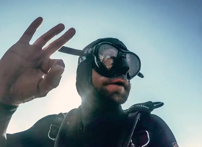 A scuba diver adjusting his goggles while holding a hand near his head in a peace sign gesture.