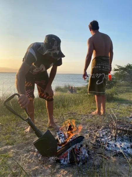 Two men on a grassy beach at sunset, one tending a small campfire with a shovel, the other standing and looking at the ocean.