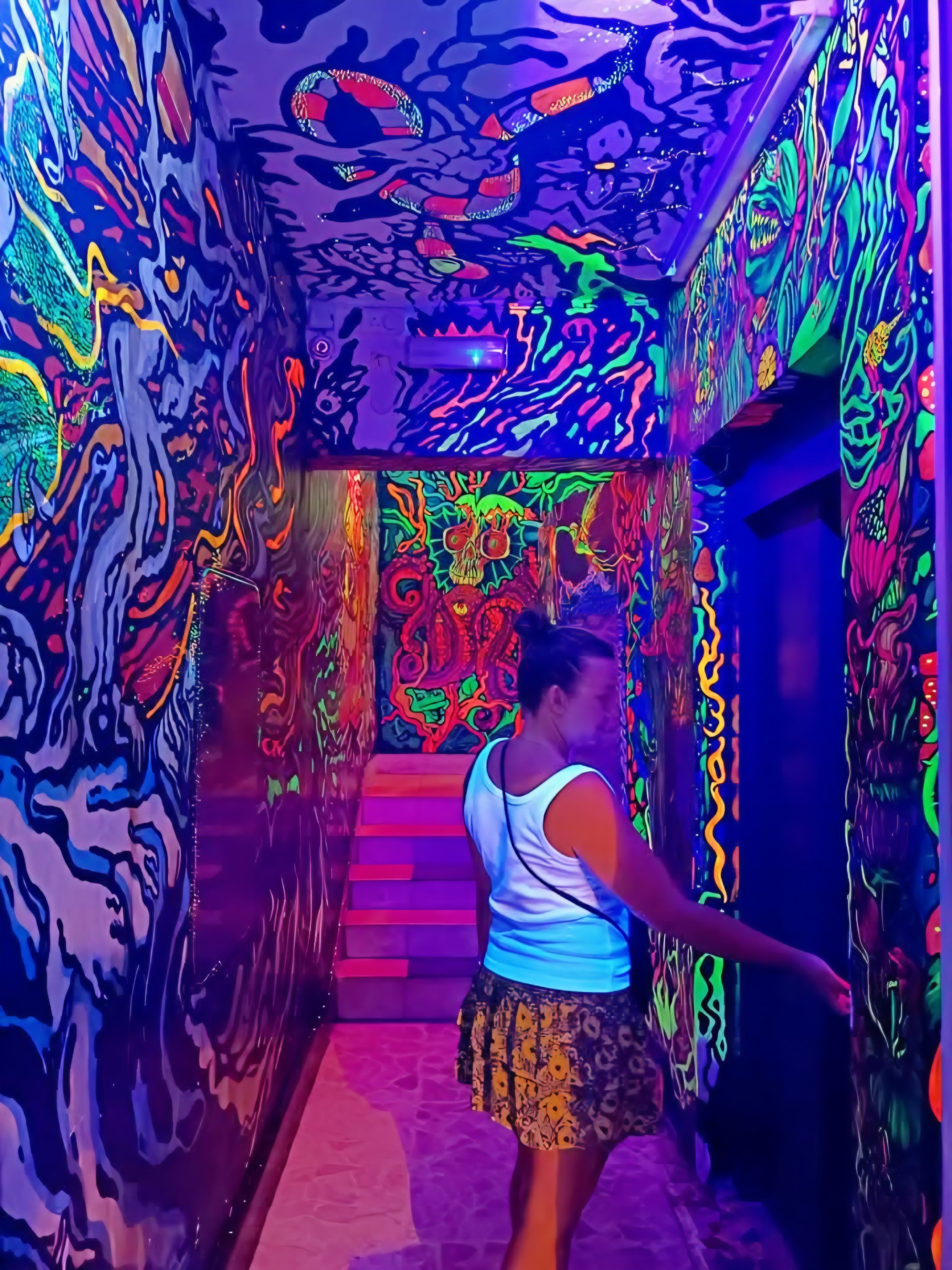 Person in a white sleeveless top and patterned shorts walking through a vibrant, neon-lit hallway with abstract, colorful murals on the walls and ceiling.