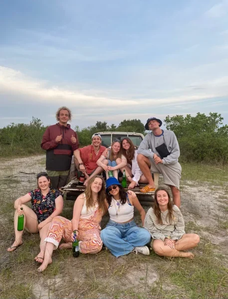 Group of eight friends outdoors near a vehicle, smiling and sitting on the ground, with a blue sky overhead.