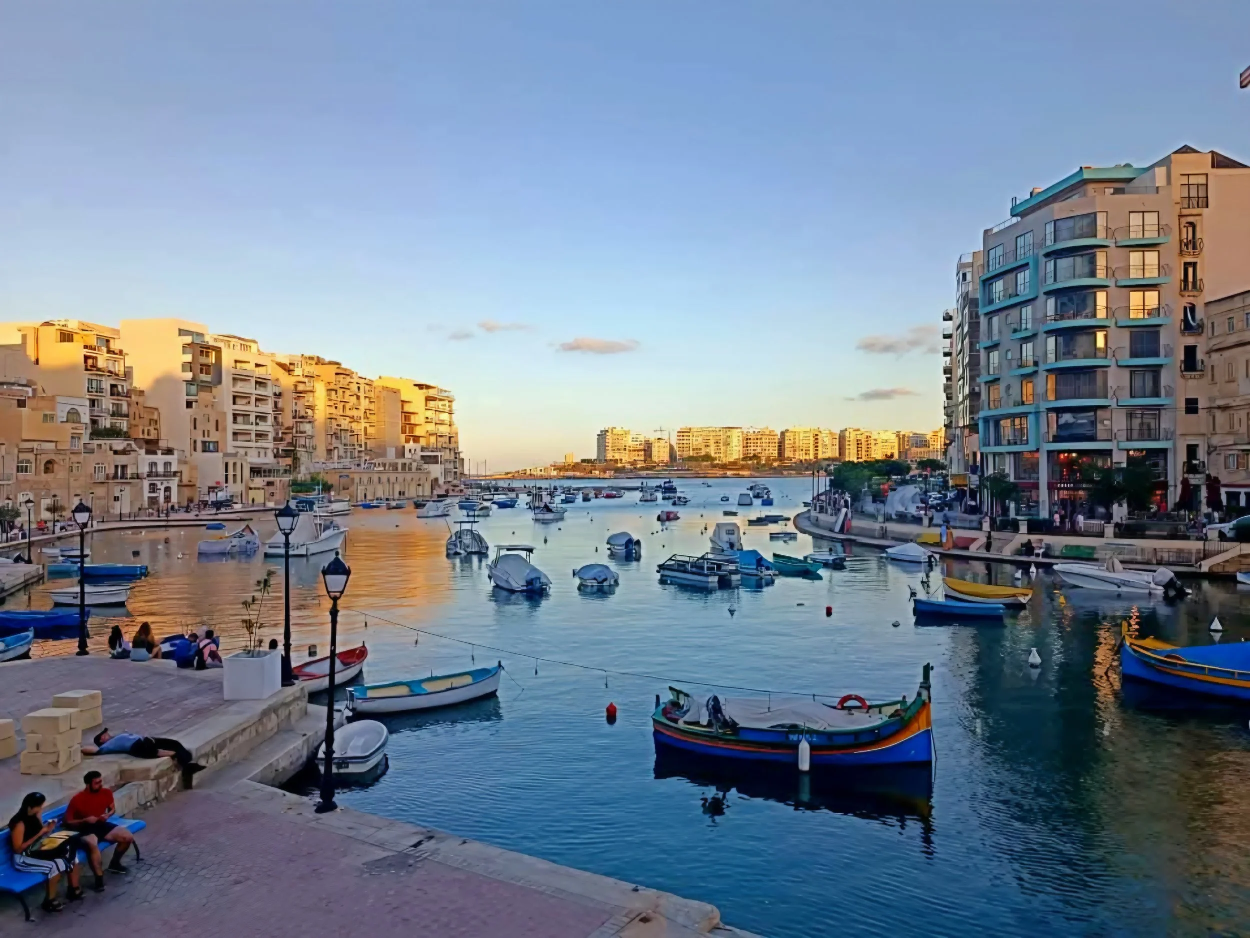 A harbor with boats docked in calm water, modern apartments on the right, and older buildings on the left, during sunset.