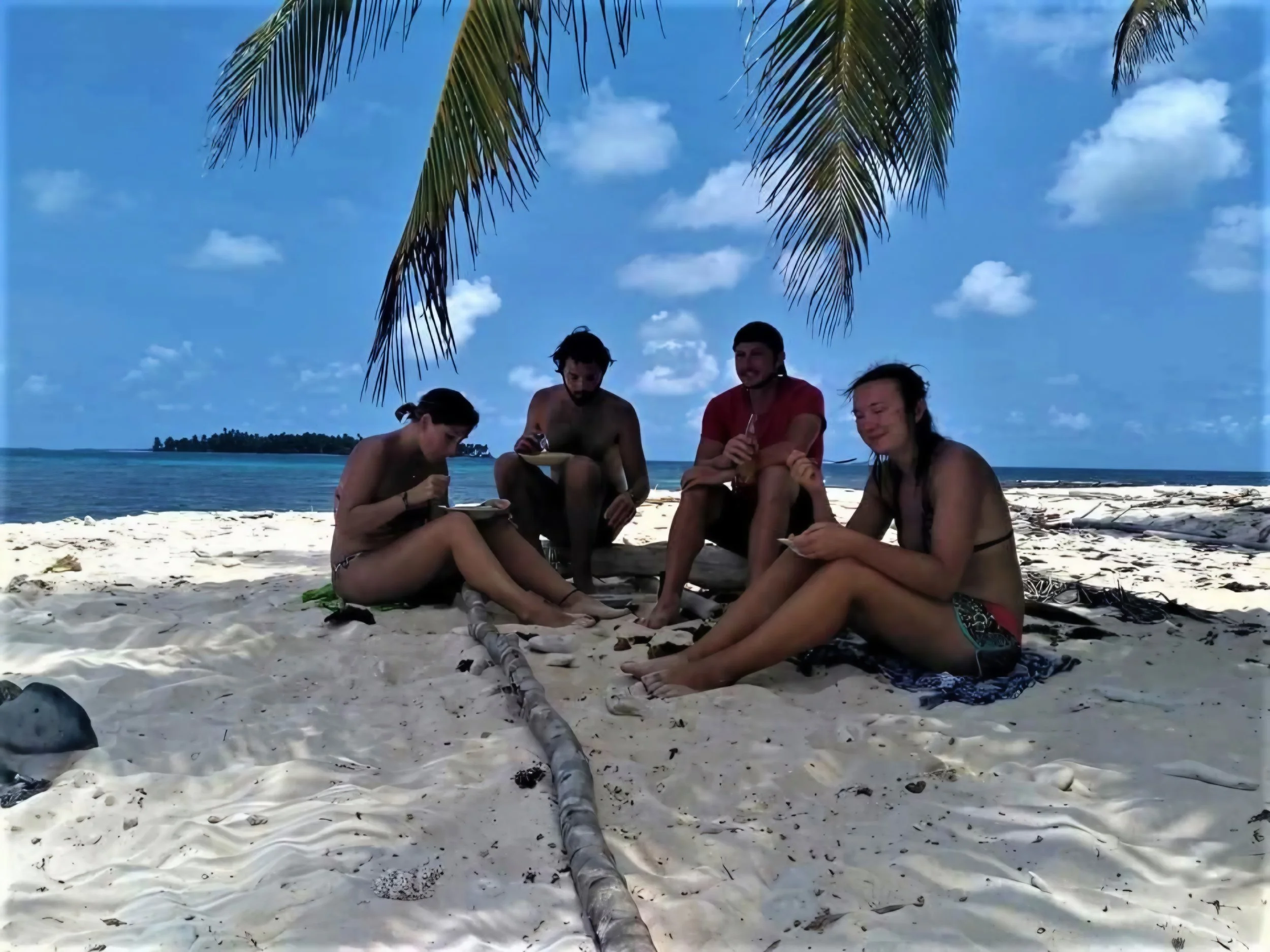 Group of five people sitting on the white sandy beach under palm trees with blue sky and clouds, looking at their phones and enjoying a sunny day.